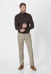 Brauner Rollkragenpullover, beige Hose und braune Schuhe. Slim Fit, gerippte Bündchen und Saum, glatte Textur. Stehende Pose mit verschränkten Armen.
