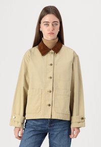 Giacca di tela beige con colletto in velluto a coste marrone, dotata di chiusura frontale con bottoni, due tasche laterali e polsini con regolazione a scatto.
