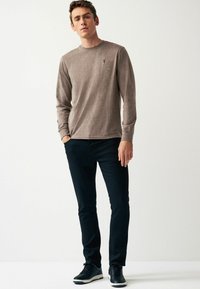 Braunes Langarmshirt mit runder Halsöffnung, kombiniert mit dunklen Slim-Fit-Hosen und schwarzen Sneakern, die ein dezentes Logo auf dem Shirt tragen.