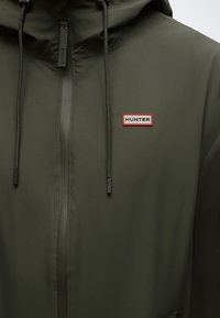 Jachetă impermeabilă verde închis cu fermoar frontal, șnururi reglabile la glugă și un logo dreptunghiular alb cu margine roșie "HUNTER" pe piept.
