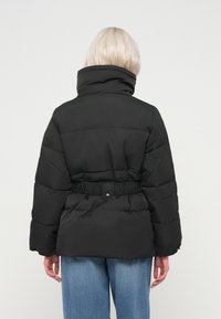 Άτομο με ανοιχτόχρωμα μαλλιά, φορώντας ένα μαύρο τζάκετ puffer με ψηλό γιακά και ελαστική μέση, συνδυασμένο με μπλε τζιν, απεικονίζεται από πίσω.