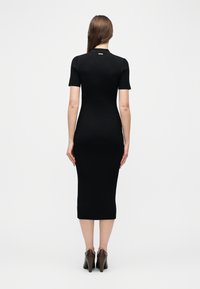 Γυναίκα που στέκεται πλάτη, φοράει εφαρμοστό μαύρο μίντι φόρεμα με ριγέ υφή, κοντά μανίκια και καφέ ψηλοτάκουνες γόβες.