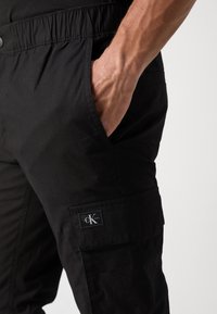 Schwarze Cargo-Hosen mit einem elastischen Bund, seitlichen Taschen und einem Patch-Logo am linken Oberschenkel, hergestellt aus einem strukturierten Stoff.