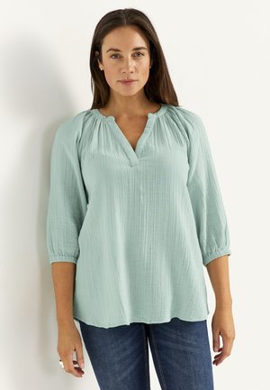 Kvinde står mod en hvid baggrund iført en lyseblå tekstureret bluse med trekvartærmer og mørkeblå jeans.