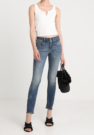 Top corto blanco acanalado con un escote en forma de ojo de cerradura, jeans ajustados azules con detalles desgastados y bolso de peluche negro sostenido en una mano.
