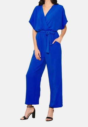 Femme portant une combinaison bleu vif à jambes larges, manches courtes, col en V, ceinture nouée à la taille, poches avant et talons ouverts noirs.