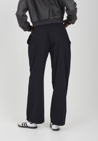 Persona che indossa pantaloni neri a gamba larga, giacca di pelle nera e sneakers bianche con strisce nere, in piedi con le mani in tasca su uno sfondo bianco.