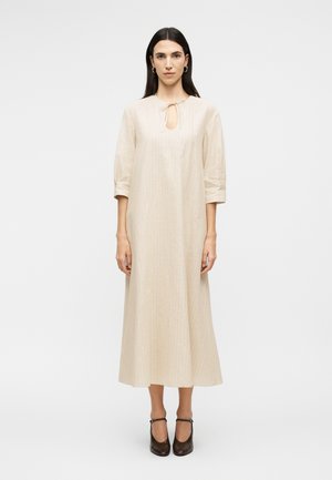 Woman with long dark hair wearing a beige long-sleeve midi dress with front tie keyhole and black Mary Jane shoes, standing on a white background.