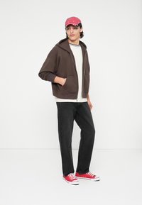 Veste à zip marron sur une chemise blanche, jeans noirs et baskets rouges. Le modèle porte une casquette rouge et noire. Design simple et coupe décontractée.