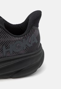 Scarpa sportiva nera con tomaia in mesh testurizzato, dotata di una spessa suola nera e logo "HOKA" in rilievo sul lato.