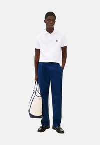 Polo bianco con un piccolo logo, abbinato a pantaloni blu navy. Tiene una borsa a tracolla beige e blu navy. Scarpe nere completano il look.