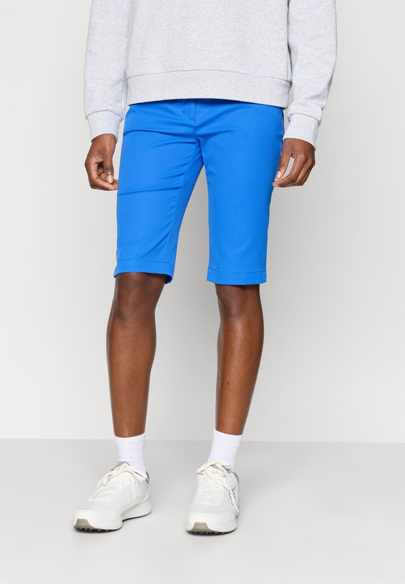 Mann iført lyseblå shorts til over kneet, hvite ankelstrømper, hvite sneakers og en lys grå hettegenser, står foran en ensfarget bakgrunn.