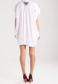 Tunic bianca, ampia e con maniche corte, con un design a farfalla all'altezza del collo sul retro. Abbinata a scarpe rosse con tacco alto.