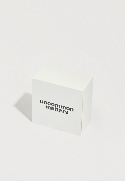 Λευκό κουτί με ματ φινίρισμα, με μαύρο κείμενο «uncommon matters» σε μία πλευρά. Τετράγωνο σχήμα, λιτός σχεδιασμός, χωρίς επιπλέον γραφικά.