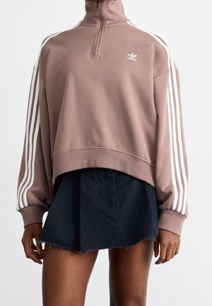Mujer con sudadera Adidas de color malva con cremallera hasta el cuello y rayas blancas en las mangas, y una falda corta negra con dobladillo sin rematar.