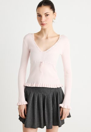 Világos rózsaszín bordázott pulóver V-nyakkal, fodros szegéllyel és hosszú ujjakkal. Sötétszürke tűcsíkos, pliszírozott szoknyával párosítva.