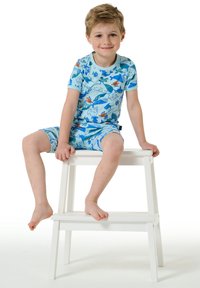 Pigiama blu a maniche corte per bambini con stampe di animali marini colorati. Il bambino è seduto su uno sgabello di legno bianco, a piedi nudi.