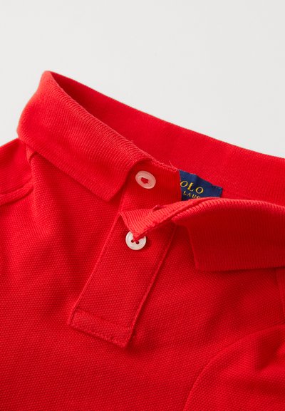 Sarkans polo krekls ar ribotu apkakli, diviem pogu slēgumiem un teksturētu audumu. Uz tā ir zils birku ar uzrakstu "Polo" dzeltenā krāsā.