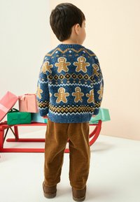 Der gestrickte Pullover hat einen blauen Grundton mit Lebkuchenmann-Mustern, strukturierte gerippte Bündchen und einen runden Ausschnitt. Kombiniert mit braunen Cordhosen.