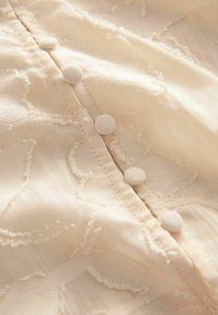 Gros plan d'un tissu beige clair avec un motif floral texturé et une rangée de boutons ronds recouverts de tissu cousus le long d'une couture.