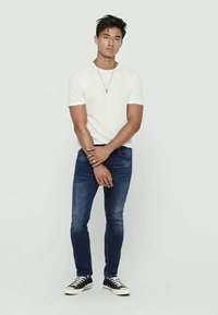 Bela majica s kratkimi rokavi, modri jeans slim fit in črne teniske. Model stoji s prekriženimi rokami, minimalni dodatki so vidni.