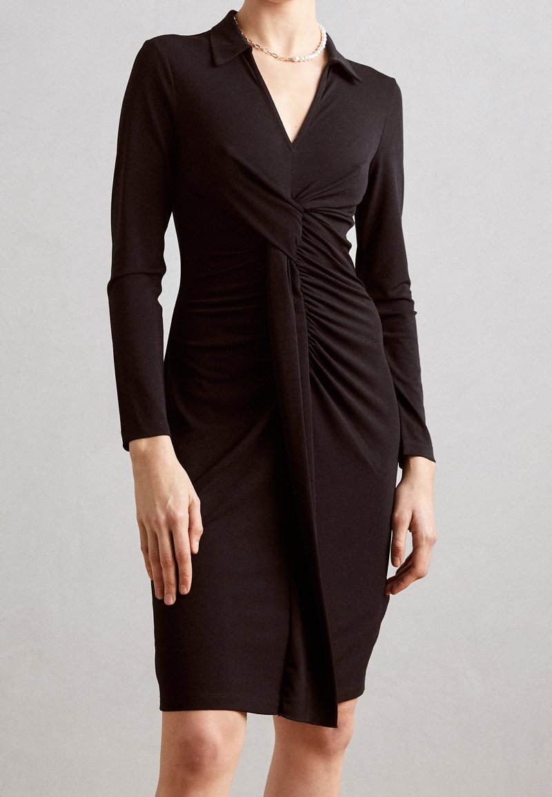 Femme portant une robe noire mi-longue à manches longues avec un détail de nœud torsadé à la taille et un col en V, debout devant un mur uni.