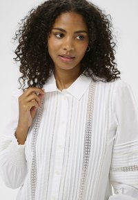 Femme aux cheveux bouclés portant un chemisier blanc avec des panneaux en dentelle verticale et des poignets smockés, regardant légèrement sur le côté, main levant près du col.