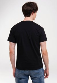 Schwarzes Baumwoll-T-Shirt mit kurzen Ärmeln, Rundhalsausschnitt und schlichten Design; getragen mit blauen Jeans. Der Stoff wirkt glatt.