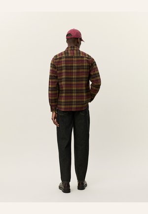 Plaid shirt in burgundy, yellow, and green tones, with a relaxed fit. Paired with dark, tapered pants and brown shoes. Cap in matching color.