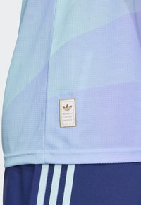 Lyseblå atletisk skjorte med strukturstof, der har et guldmærke, som siger "Authentic Licensed Product." Mønstrede med blød gradient.