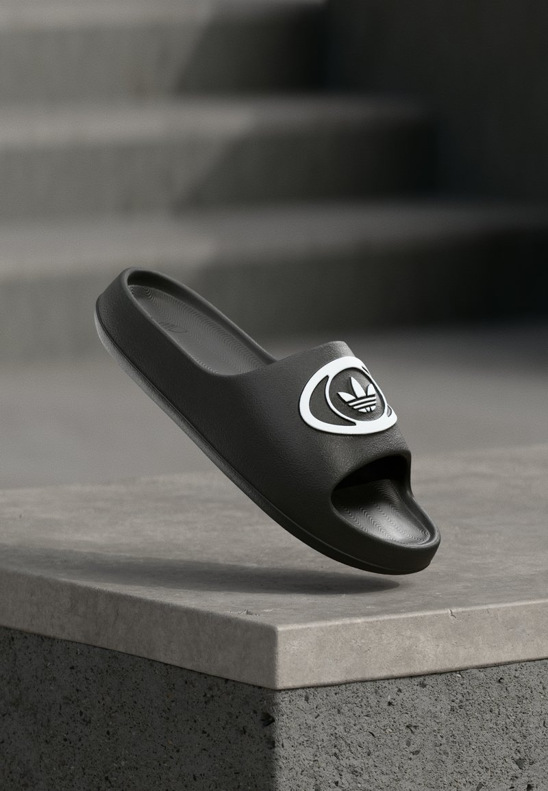 Crna natikača s otvorenim prstima, s glatkim sintetičkim gornjim dijelom i bijelim logotipom. Teksturirana potplat i zaobljen dizajn.