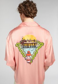 Camicetta in satin rosa a maniche corte con una grafica colorata sul retro che raffigura un tramonto, un molo e una figura che indossa un cappello.