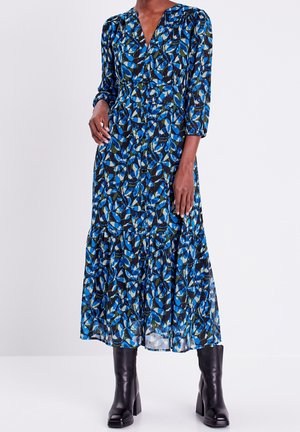 Femme portant une robe à manches longues avec des boutons sur le devant et un motif de feuilles bleues et vertes, associée à des bottines noires à talons, debout sur fond blanc.