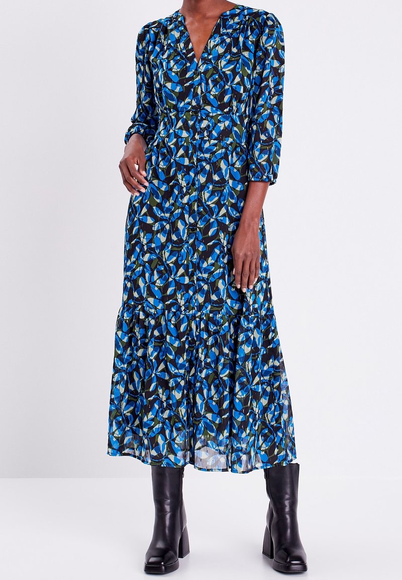 Femme portant une robe à manches longues avec des boutons sur le devant et un motif de feuilles bleues et vertes, associée à des bottines noires à talons, debout sur fond blanc.