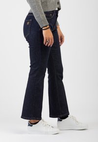 Jeans in denim a gamba larga di colore blu scuro con cuciture arancioni a contrasto, tasche laterali e un logo visibile sul retro. Abbinati a sneakers bianche.