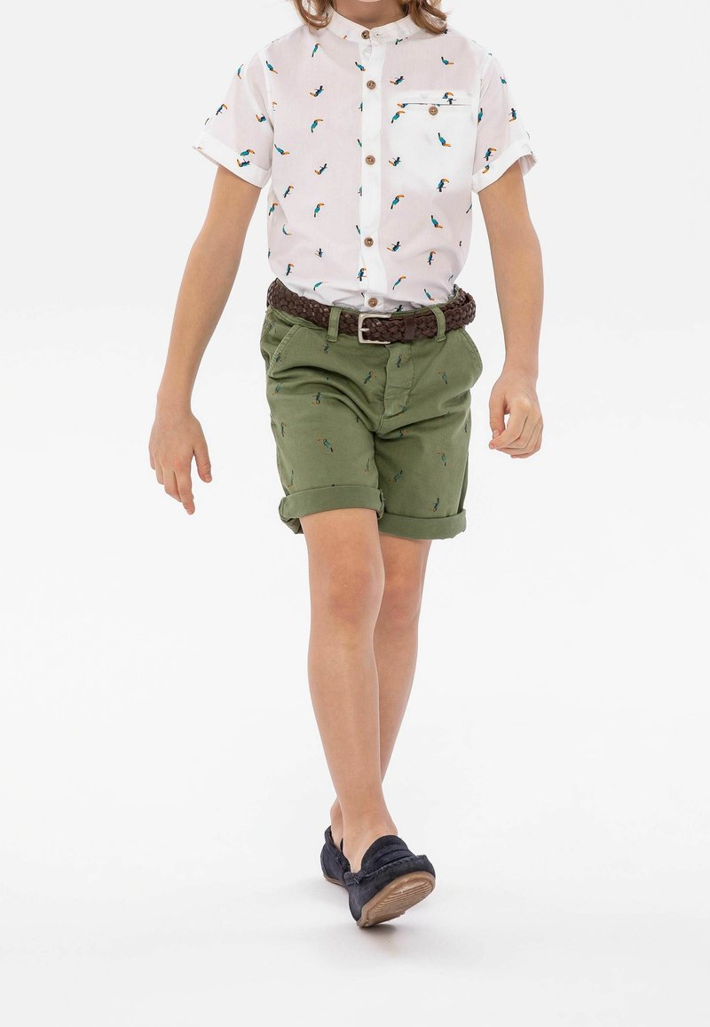 Enfant marchant, portant une chemise blanche à manches courtes avec des imprimés d'oiseaux, un short vert retroussé, une ceinture tressée marron et des mocassins noirs.