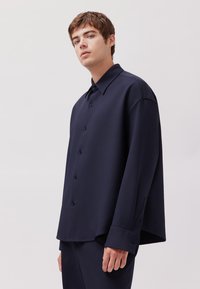 Camicia blu navy con bottoni, dal taglio rilassato, caratterizzata da un colletto aperto, maniche lunghe e una texture di tessuto liscia.