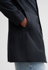 Navy blue coat with a smooth texture, single button closure, and a side pocket. Worn over light blue jeans.