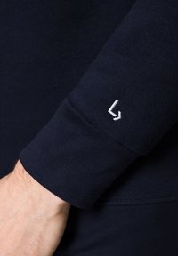 Granatowo niebieska bawełniana odzież z długim rękawem i dopasowanym mankietem, ozdobiona małym białym haftowanym logo na rękawie. Gładka tekstura.