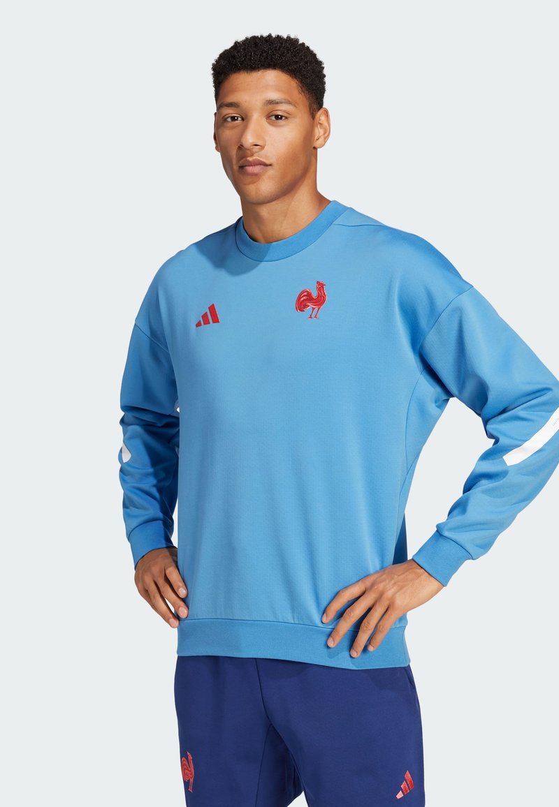 Sininen huppari, jossa on pitkät hihat, brodeerattu punainen kukko-logo ja Adidas-brändäys. Valmistettu pehmeästä kankaasta, jossa on pyöreä pääntie.
