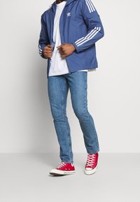Blå lättviktsjacka med vita ränder, vit t-shirt, blå jeans och röda hög-top sneakers. Avslappnad, sportig outfit med tydlig branding.