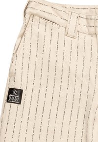 Beige pants featuring vertical printed text patterns. Includes a black label at the waistband. Made from a lightweight, cotton-like material.