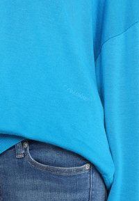 Blauer Strickpullover mit gerippter Struktur, in lässiger Oversize-Passform, kombiniert mit mittelblauen Denim-Jeans.