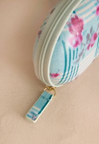 Vetoketjullinen sylinterimäinen kotelo, jossa on vaaleansininen tausta ja pinkki-sininen kukkakuosi, kullanvärinen vetoketjun veto ja raitoja.