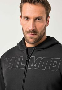 Sudadera con cremallera negra con un frente texturizado que presenta "UNLMTD" en letras gruesas, fabricada en un tejido suave y con capucha de cuello redondeado.