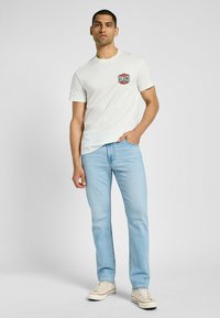 Biała bawełniana koszulka z czerwono-niebieską naszywką z logo "Lee Jeans", zestawiona z jasnoniebieskimi dżinsami i beżowymi sneakersami.