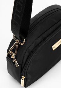 Černá nylonová taška přes rameno s hladkou strukturou, zlatým zipem a kovovými detaily. Obsahuje štítek s logem a široký nastavitelný popruh.