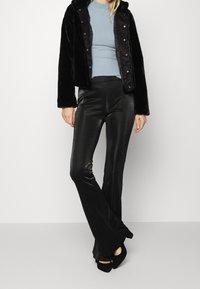 Veste en fausse fourrure noire avec fermeture pression sur le devant, haut côtelé bleu clair et pantalon noir évasé brillant. Des chaussures à talons hauts noires complètent le look.