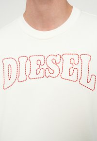 Detailný záber na biele tričko s okrúhlym výstrihom, na ktorom je cez hrudník prešitý nápis "DIESEL" červenou niťou.