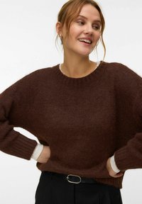 Jeune femme aux cheveux clairs portant un pull marron foncé en tissu duveteux par-dessus un t-shirt blanc à manches longues et un pantalon noir avec une ceinture noire, souriante et regardant sur le côté.